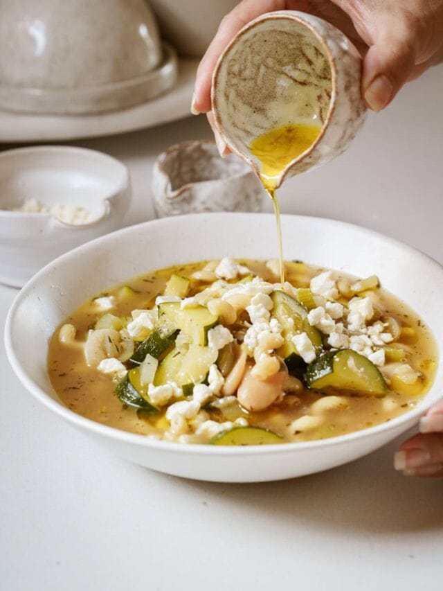 A person drizzles sauce over a bowl of vegetable and pasta soup. The soup includes zucchini, beans, and crumbled cheese. There are ceramic bowls in the background on a white surface.