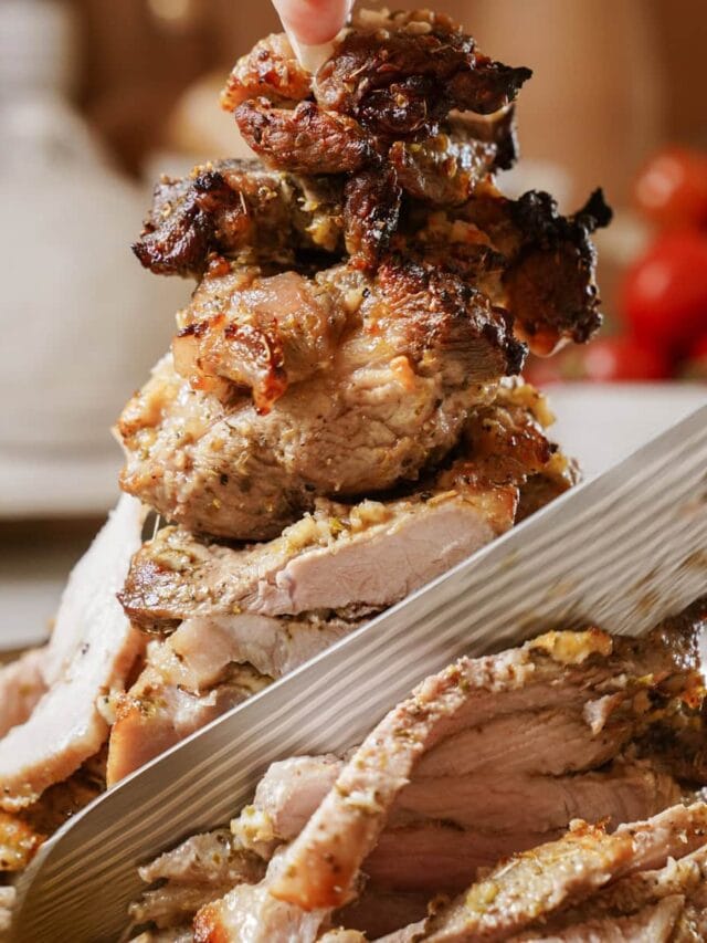 Close-up of a person slicing a stack of roasted, seasoned meat with a visible knife. The meat appears juicy and tender, with a crispy exterior.