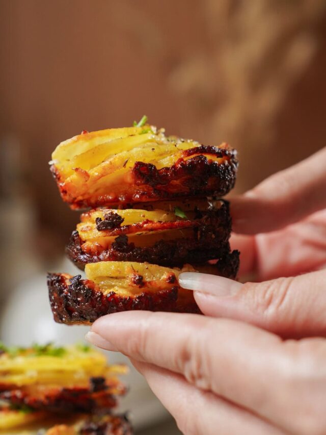 Close-up of a person holding a stack of crispy, golden-brown roasted potato slices seasoned with herbs. The potatoes are perfectly layered and look appetizing, with a slightly charred edge. A warm, blurred background completes the image.