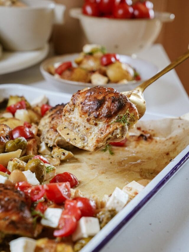 A close-up of a spoon lifting a piece of cooked chicken from a sheet pan filled with sliced potatoes, cherry tomatoes, olives, and cubes of feta cheese. In the background, a bowl of cherry tomatoes is partially visible.