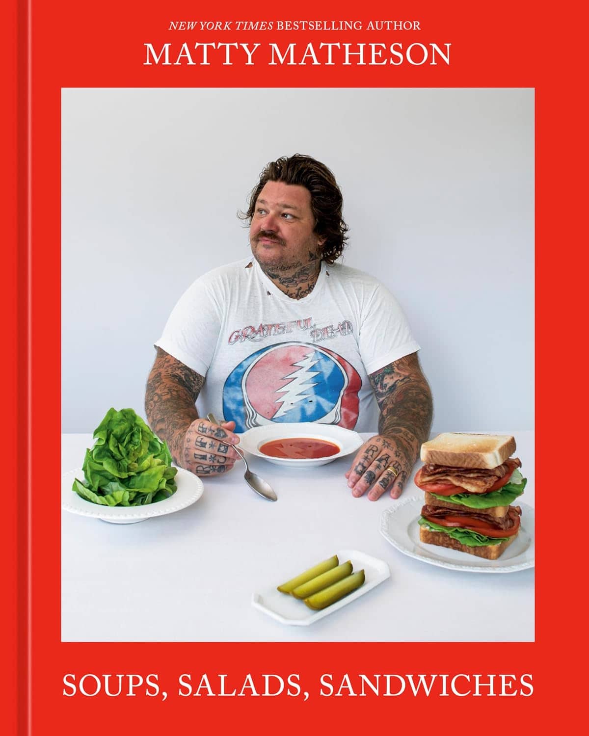 The cover of a cookbook features a man with tattoos sitting at a table with plates of lettuce, soup, a sandwich, and pickles. The title reads Soups, Salads, Sandwiches and the authors name is Matty Matheson.