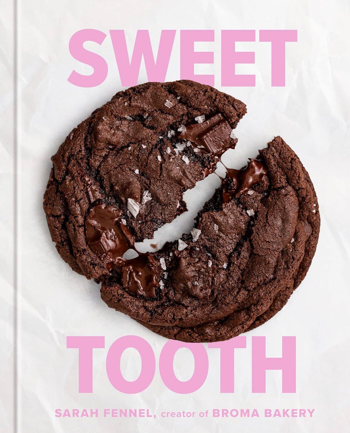 A broken chocolate cookie with chunks and a sprinkle of sea salt is centered on a white background. The words Sweet Tooth appear in large pink letters above the cookie, and the creators name is mentioned below.