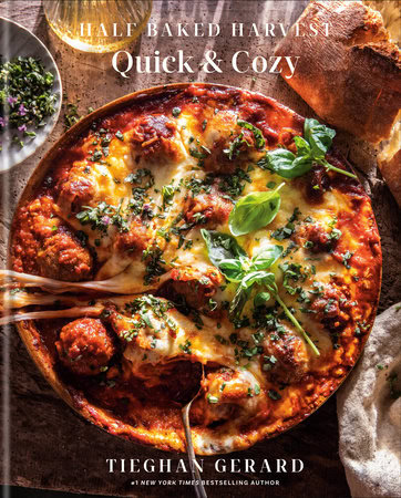 A cookbook cover titled Half Baked Harvest: Quick & Cozy by Tieghan Gerard. The image features a cheesy baked dish topped with herbs and greens, surrounded by pieces of crusty bread.