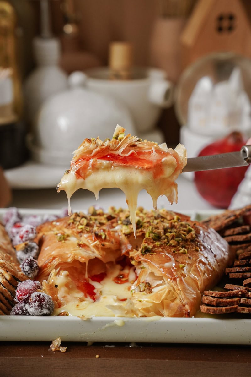 A baked pastry shell oozing with melted cheese and topped with crushed nuts is being sliced. Surrounded by crackers and berries, it sits on a white platter, invitingly set in a cozy kitchen setting.