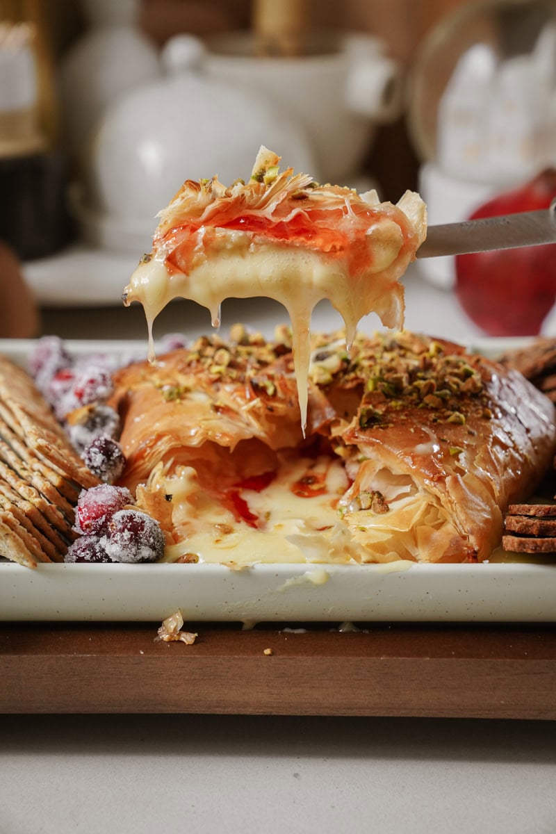 A close-up of a baked Brie wrapped in phyllo dough. The Brie is topped with pistachios and a slice is being pulled away, showing melted cheese and red jam. It is served on a platter with crackers and sugared cranberries.