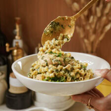 A close-up of a bowl filled with creamy pasta, chicken pieces, and kale, being served with a gold spoon. The background includes kitchen items like bottles and a faucet, with a blurred effect.