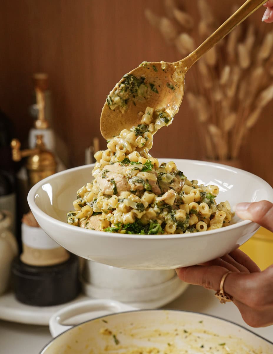 A person serves creamy pasta with herbs from a pot into a white bowl. The pasta features small noodles and is mixed with chicken and greens. The background includes kitchen decor with bottles and a dried plant arrangement.