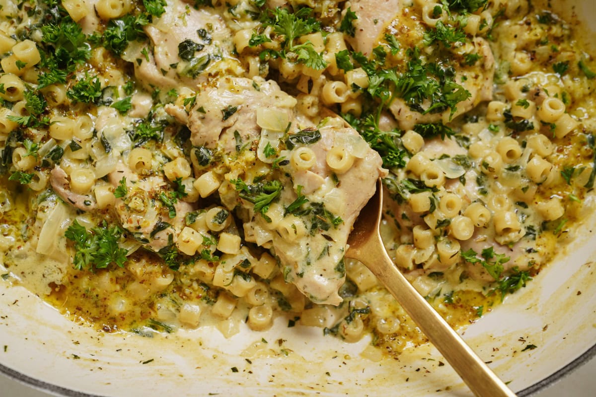 A creamy pasta dish with pieces of chicken, small pasta tubes, and a rich cream sauce. The dish is garnished with chopped parsley and appears to be freshly cooked, with a golden spoon resting in the pan.