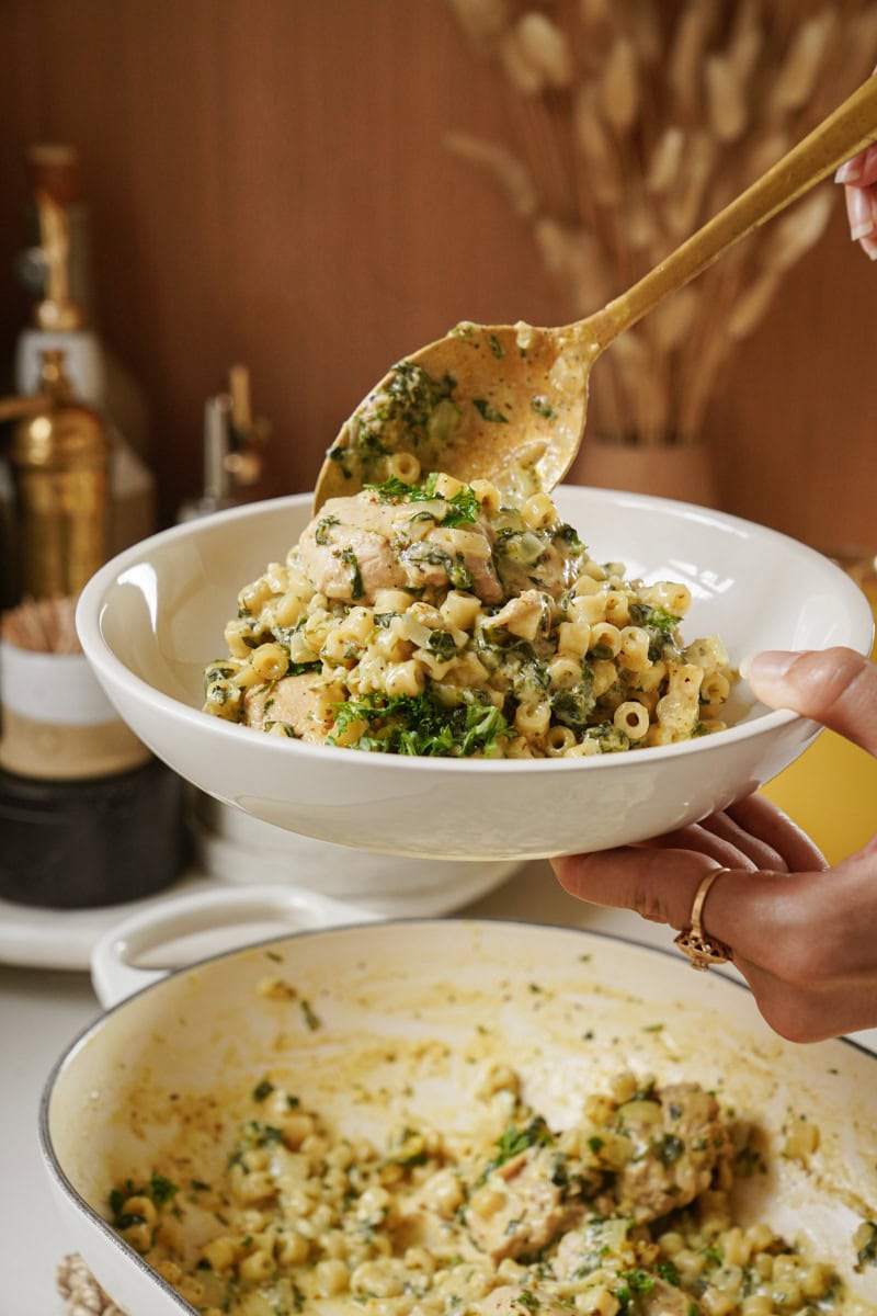 A person serving creamy pasta with greens, chicken, and herbs from a pot into a white bowl with a golden spoon. The background features kitchen items and dried plants on a wooden surface.