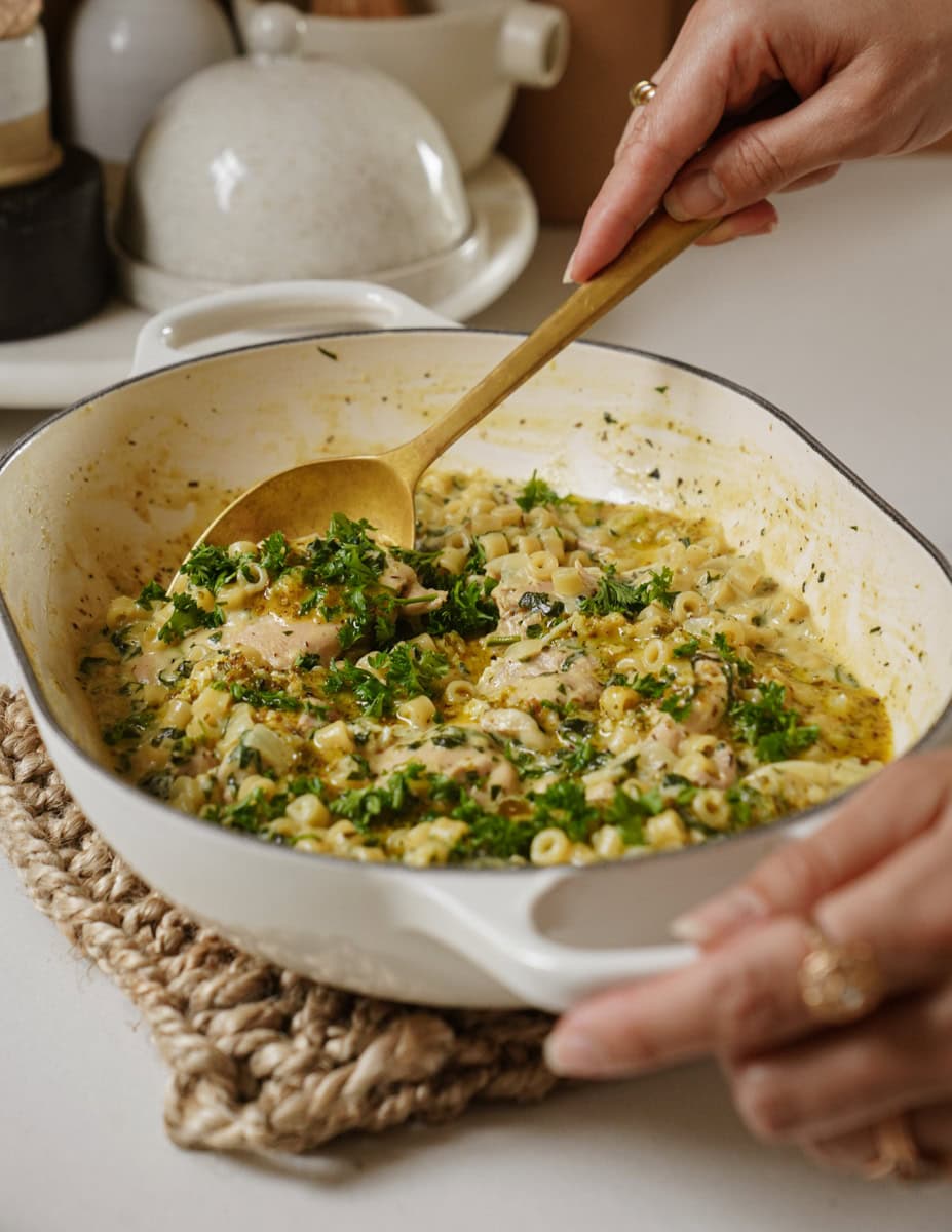 A person stirs a creamy pasta dish with chicken and finely chopped parsley in a white casserole dish. The meal rests on a woven mat on a white countertop; salt and pepper shakers are visible in the background.