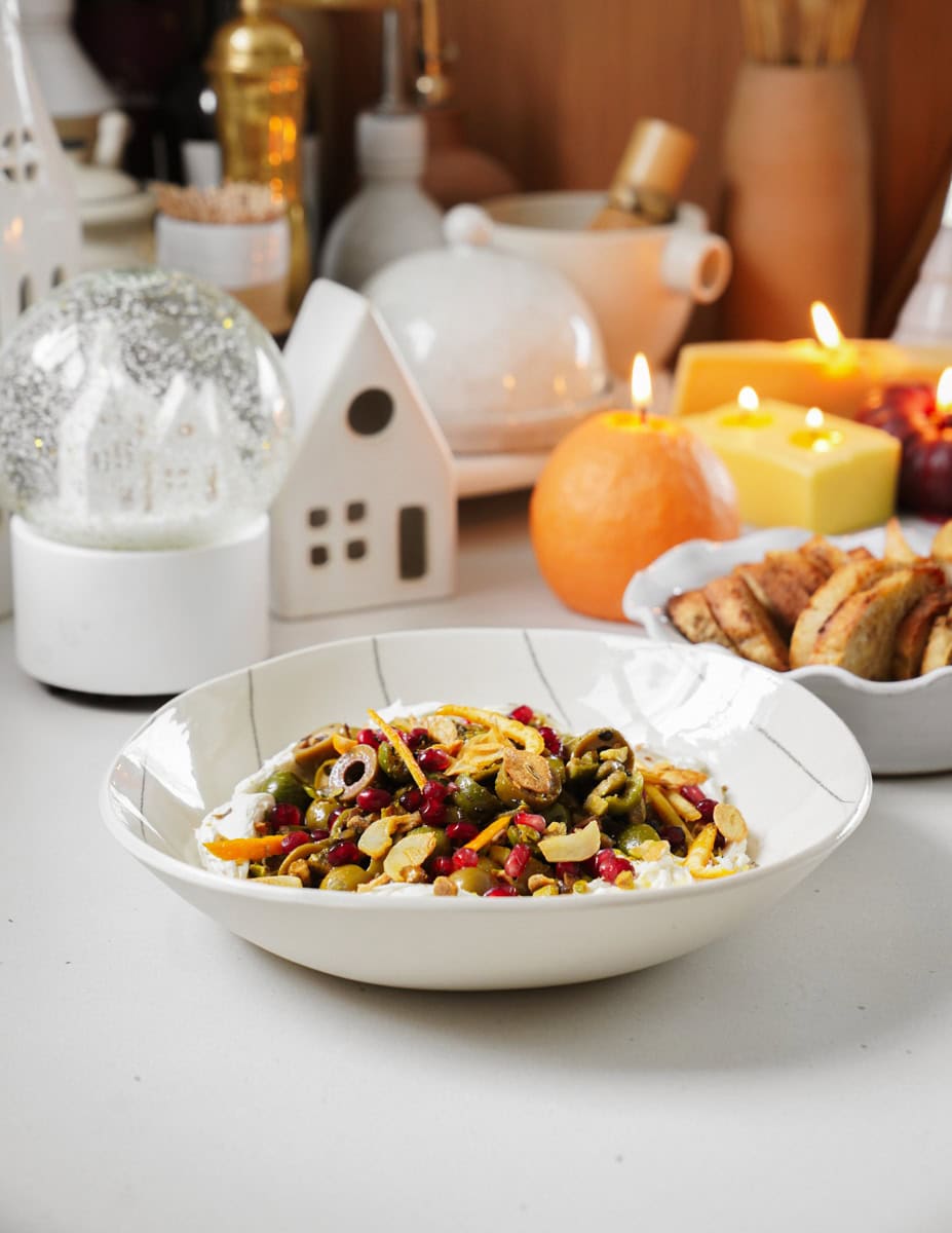 A bowl of mixed olives, pomegranate seeds, and nuts on a white plate. The background features lit candles, a snow globe, small decorative houses, and a cozy kitchen setting.