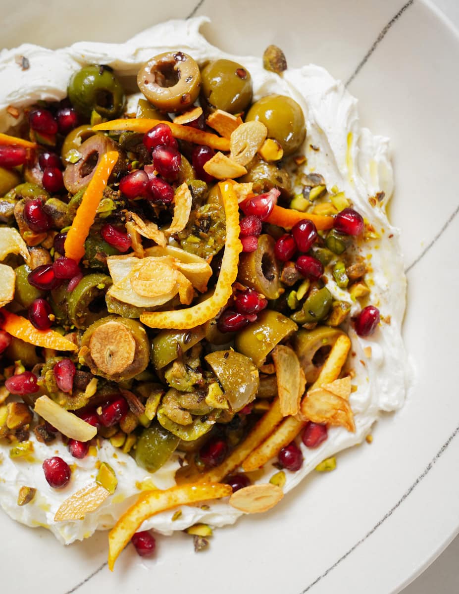 A dish featuring whipped feta topped with sliced green olives, pomegranate seeds, orange zest, pistachios, and garlic chips, served on a white plate.