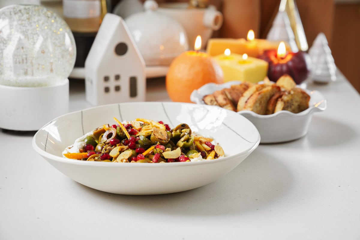 A bowl of salad featuring olives, corn, and pomegranate seeds sits on a table. In the background, there are lit candles, a white ceramic house decor, and a dish of cookies. An orange adds a pop of color beside the cookies.