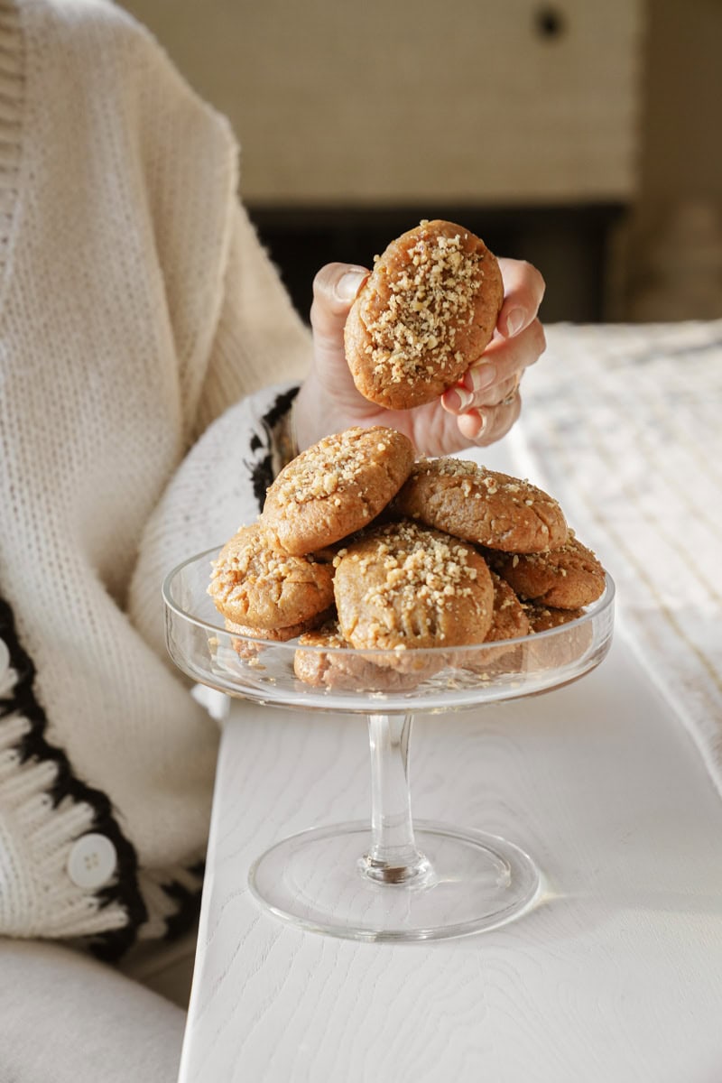 A person wearing a white sweater holds up a cookie above a dish filled with cookies topped with crushed nuts. The dish is on a white table, creating a cozy and inviting atmosphere.