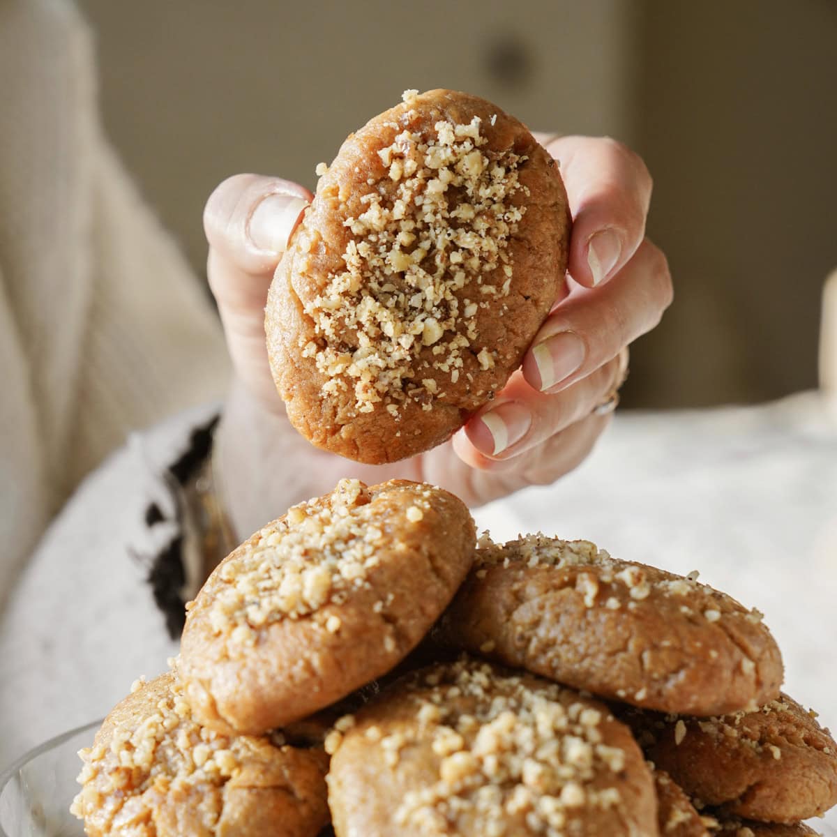 A hand holding a cookie topped with crushed nuts above a plate of similar cookies. The cookies are golden brown, textured, and appear freshly baked. The background is softly blurred, highlighting the cookies as the focal point.