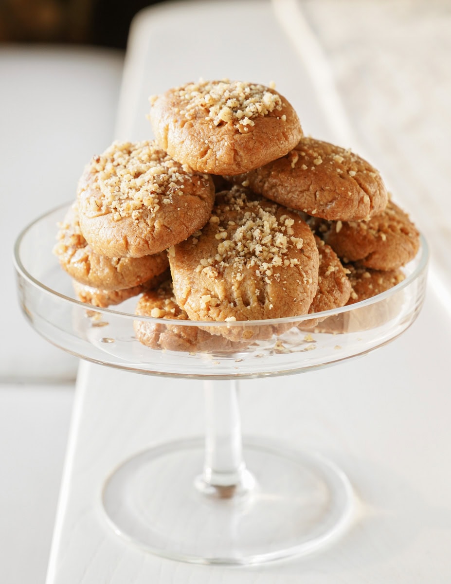 A glass pedestal dish holds a stack of round cookies sprinkled with crushed nuts. The cookies have a golden-brown color and a crumbly texture, sitting on a white surface in a softly lit setting.