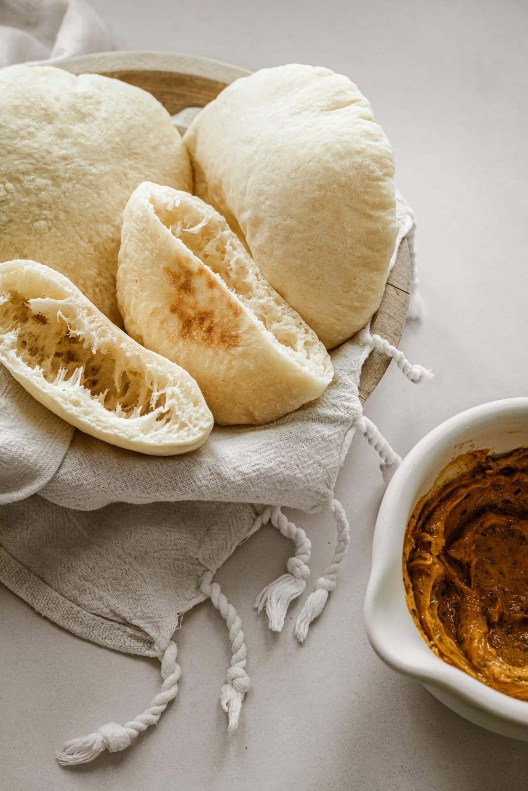 A basket of freshly baked pita bread sits on a white cloth, with one piece split open to reveal its fluffy interior. Next to the basket is a bowl containing a creamy, spiced dip.