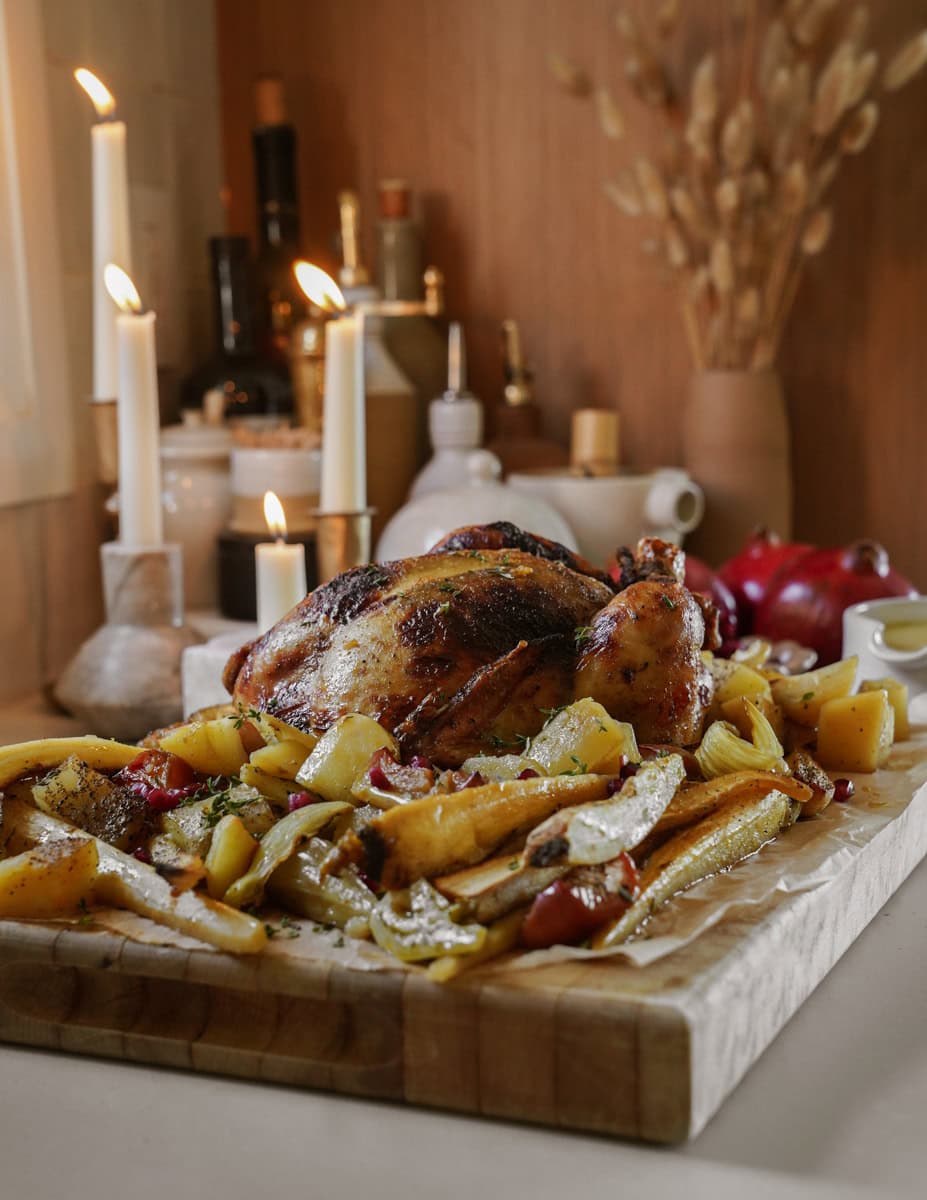 Roasted chicken and vegetables on a wooden cutting board. Candles and decorative items adorn the background, creating a cozy, rustic atmosphere.