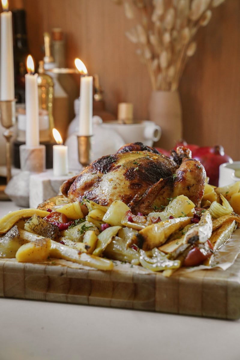 A roasted chicken on a wooden board is surrounded by roasted vegetables, including potatoes and carrots. Lit candles and dried flowers create a warm, cozy atmosphere in the background.