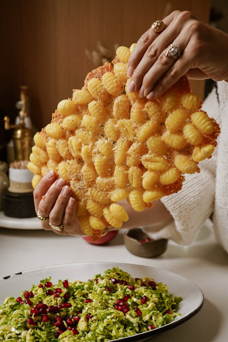 A person holding a golden, crispy layer of baked gnocchi topped with cheese and spices. Below, a plate of fresh salad with shredded greens and pomegranate seeds is on the table.