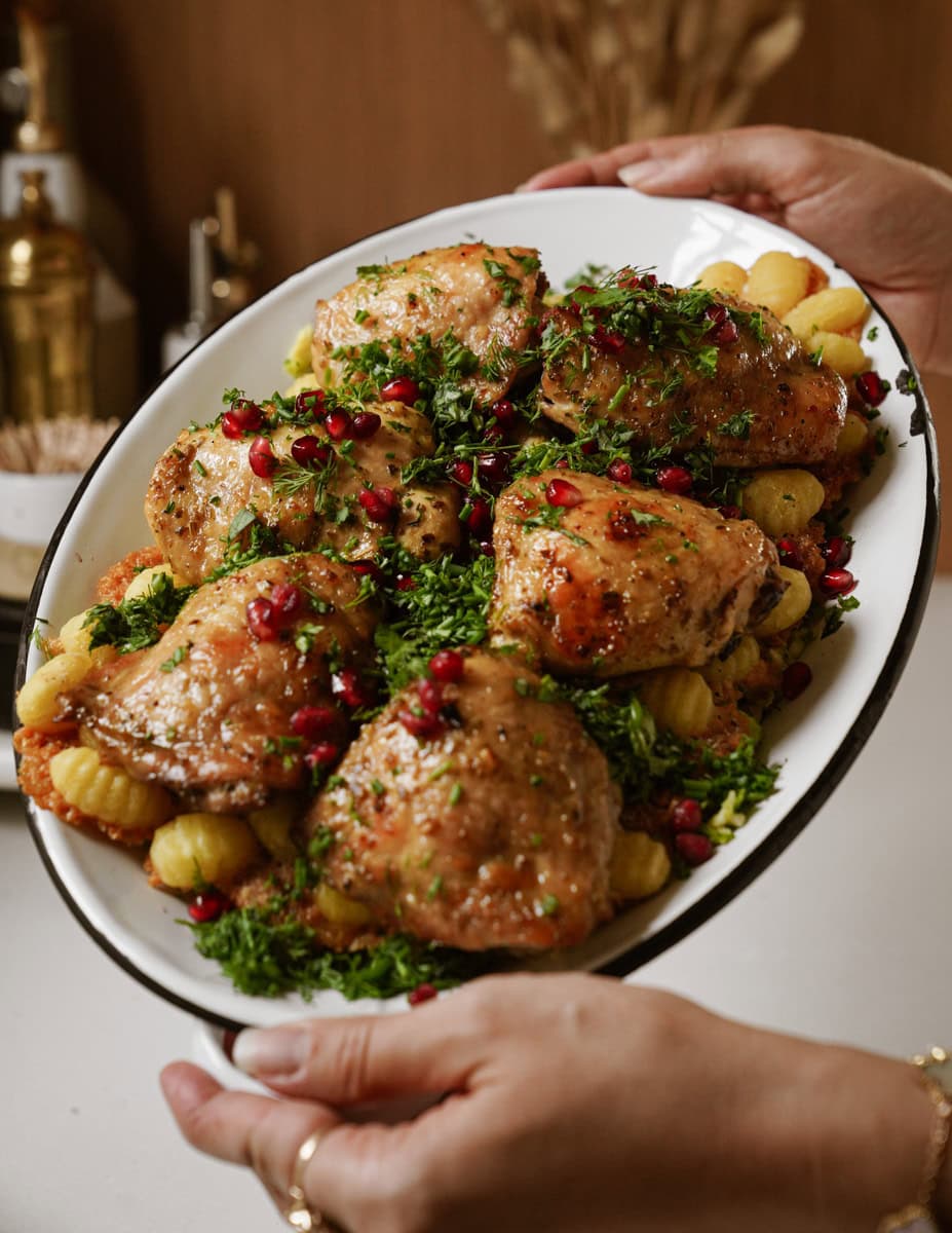 A person holds an oval platter filled with roasted chicken thighs, garnished with fresh herbs and pomegranate seeds, served over a bed of gnocchi. The dish is set against a kitchen backdrop.