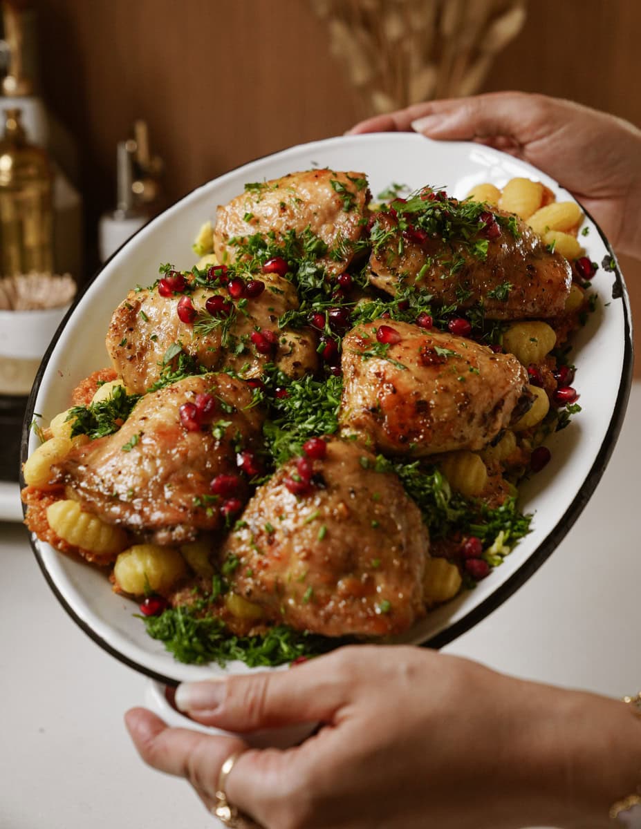 A platter of roasted chicken thighs garnished with fresh parsley and pomegranate seeds, served on top of roasted baby potatoes. Two hands are holding the dish.