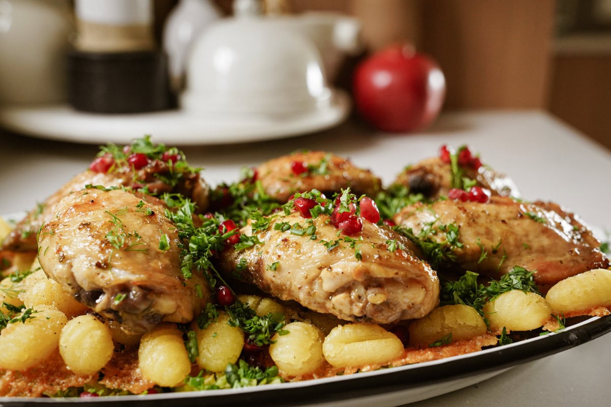 A plate of roasted chicken pieces garnished with green herbs and red pomegranate seeds, served on a bed of golden gnocchi.