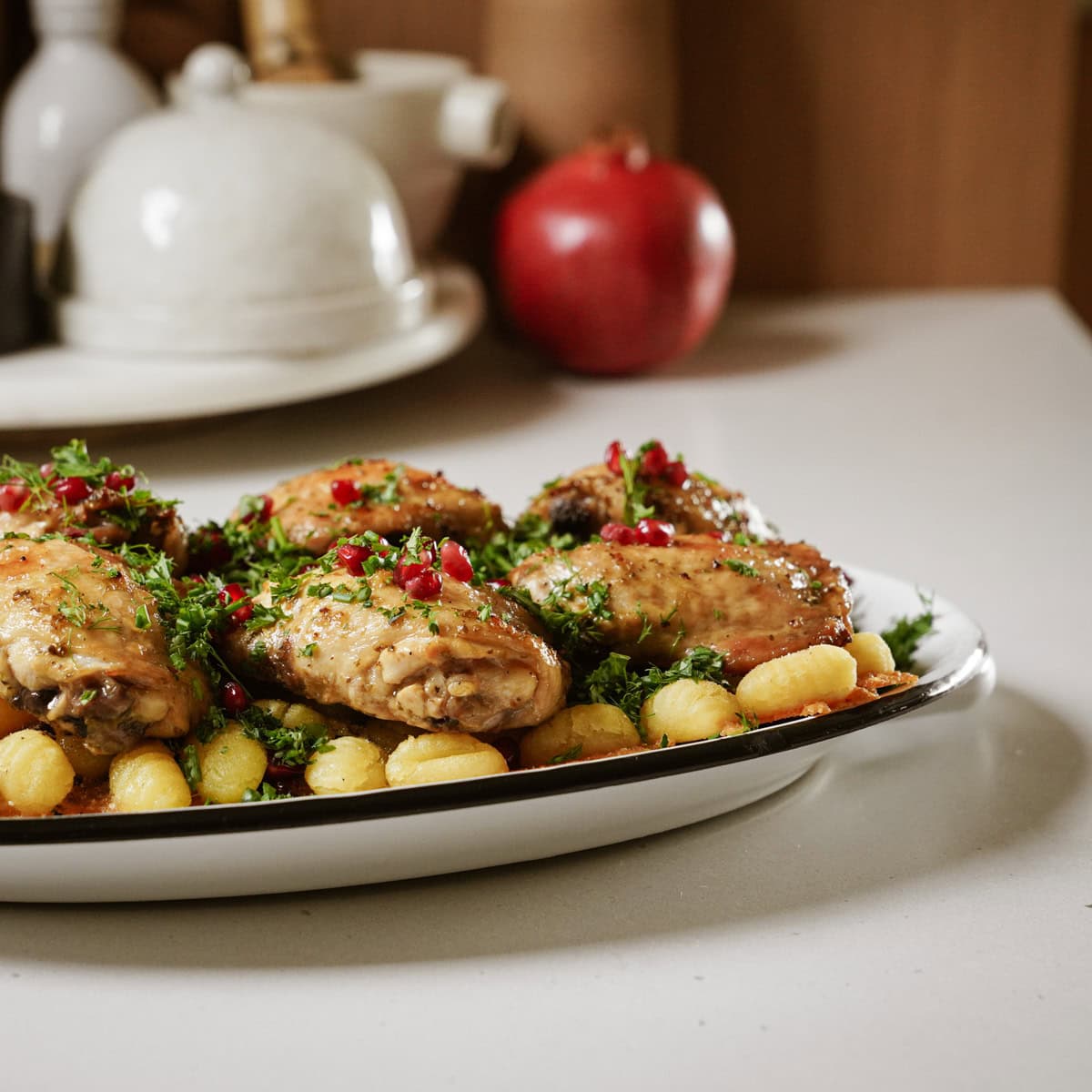 A plate of roasted chicken thighs garnished with red seeds and herbs, served on sheet pan gnocchi. In the background, a pomegranate and a kitchen item are partially visible on a countertop.