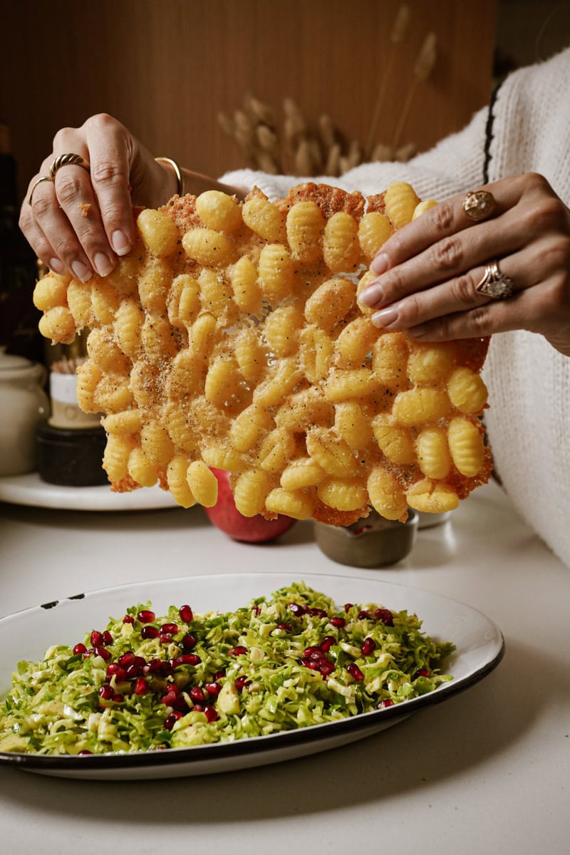 A person holding up a large, crispy fried gnocchi sheet over a plate of chopped green salad with pomegranate seeds. The background shows a kitchen setting with jars and utensils.