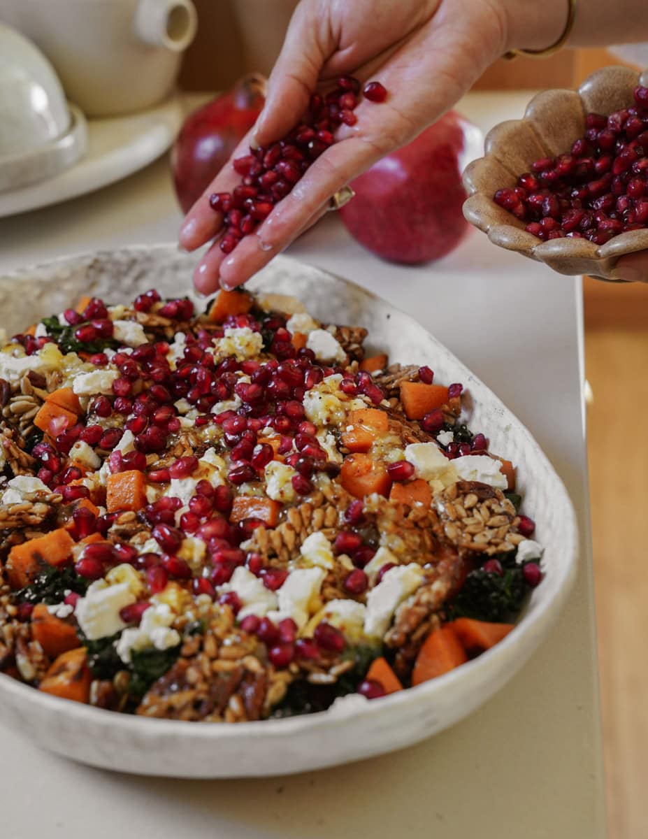 A person is sprinkling pomegranate seeds onto a salad in a white dish. The salad contains pieces of squash, kale, nuts, and crumbled cheese. Two whole pomegranates are visible in the background.