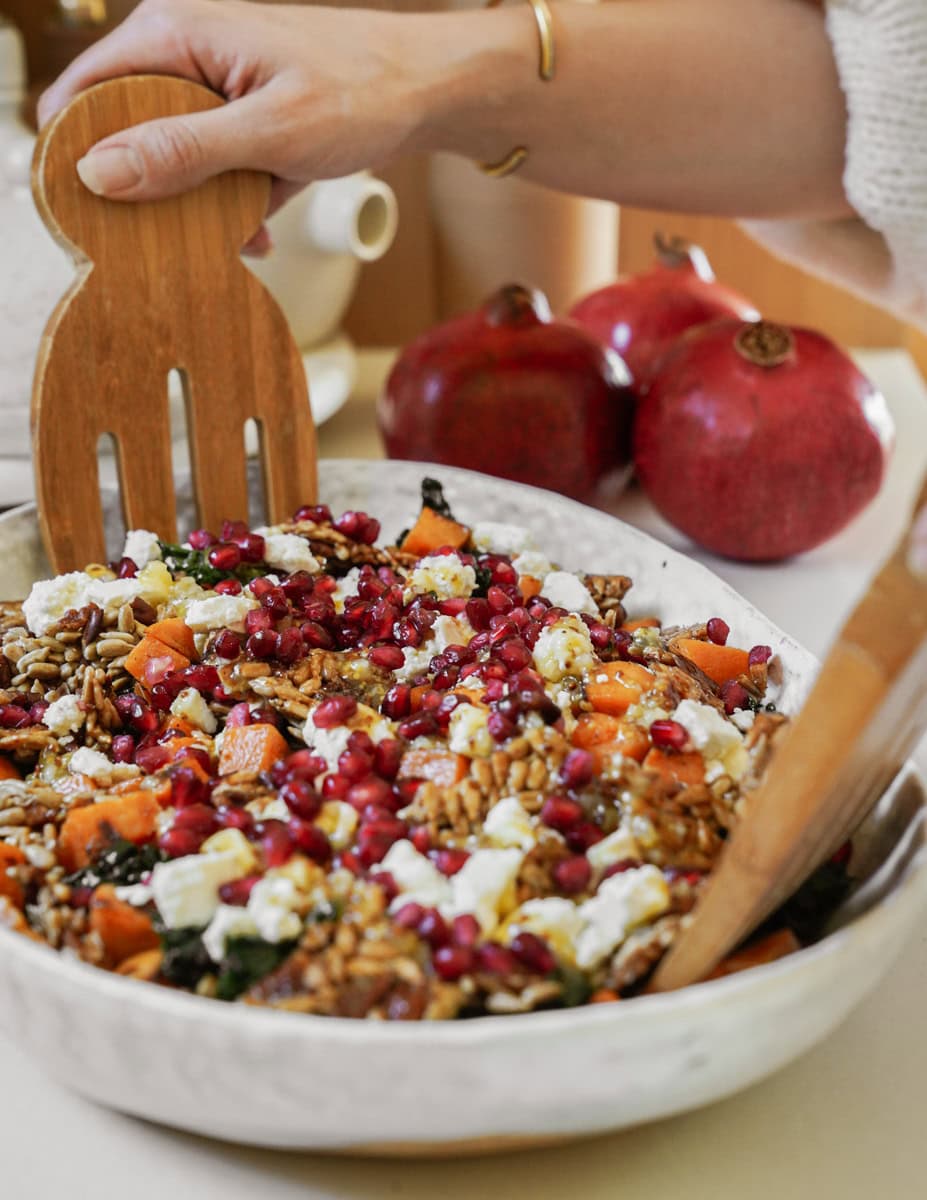 A person tosses a colorful salad with a wooden fork in a bowl. The salad includes grains, pomegranate seeds, feta cheese, leafy greens, and roasted squash. Two pomegranates are visible in the background.