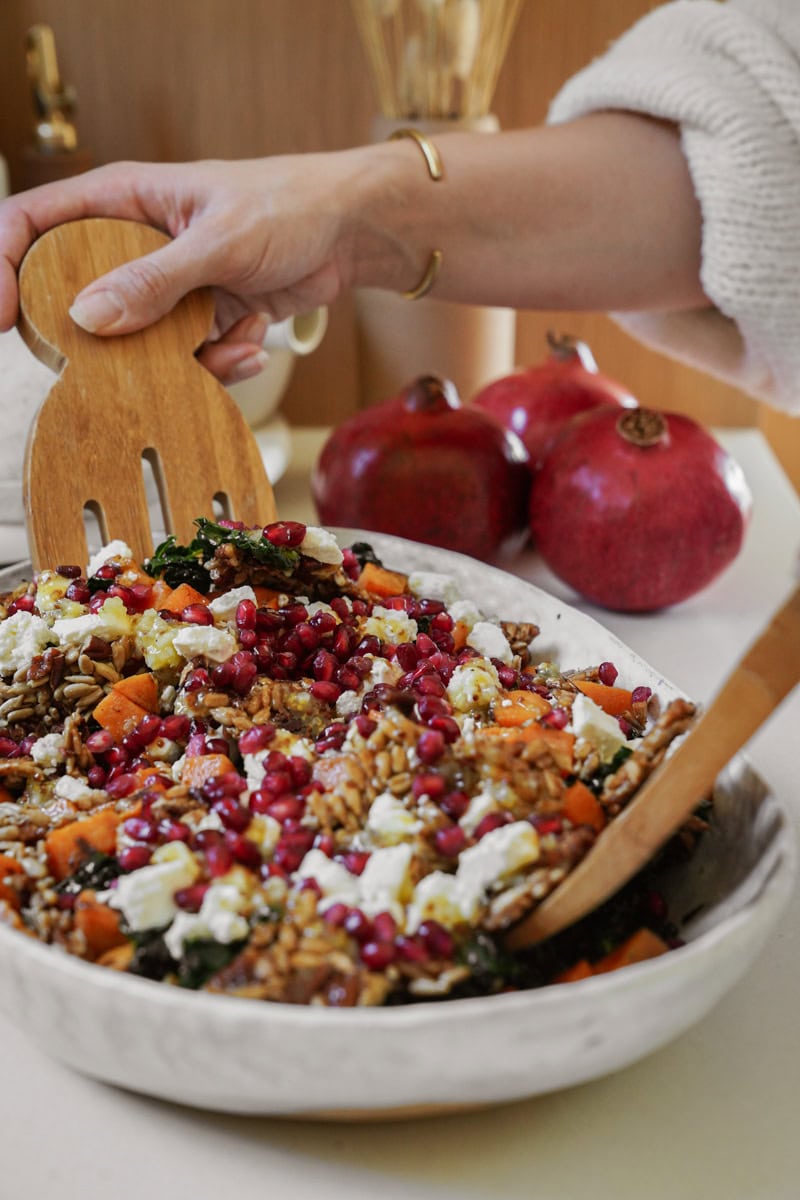 A hand using wooden utensils to serve a salad from a bowl. The salad includes grains, roasted vegetables, crumbled cheese, and pomegranate seeds. Three pomegranates are in the background on a countertop.