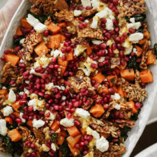 A close-up of a colorful salad featuring sweet potatoes, crumbled feta, pomegranate seeds, kale, and crunchy seeds or nuts, all drizzled with a light dressing. The dish is served in an oval platter.