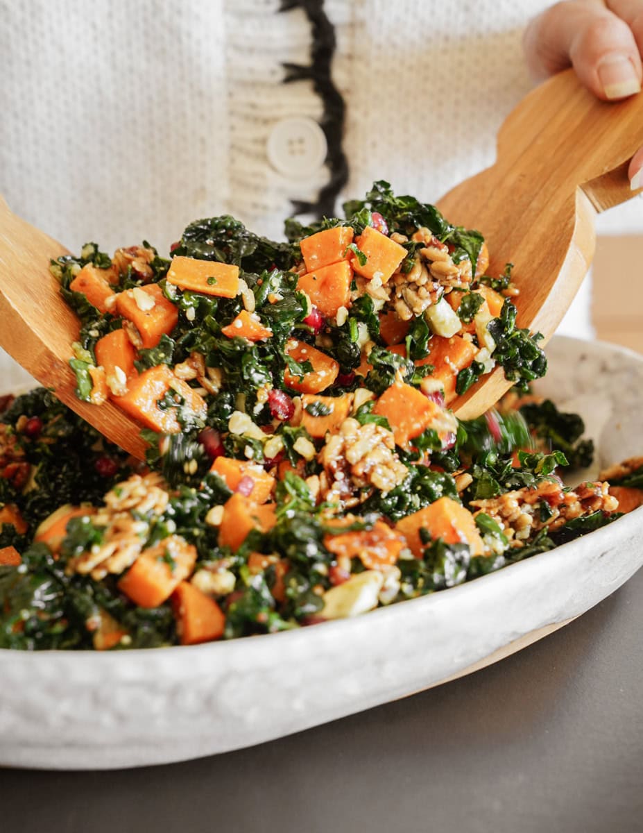 A person tosses a colorful salad with wooden salad tongs in a white bowl. The salad includes kale, diced orange squash, seeds, nuts, and pomegranate seeds. The person is wearing a white sweater with a visible button.