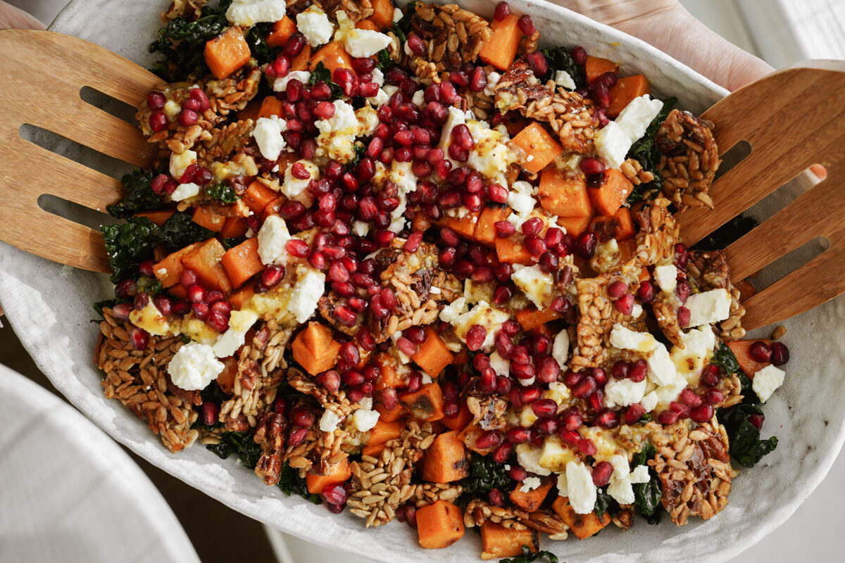 A vibrant salad with roasted sweet potatoes, pomegranate seeds, feta cheese, and sunflower seeds, served on a white platter. Large wooden salad tongs are positioned at the top of the dish.