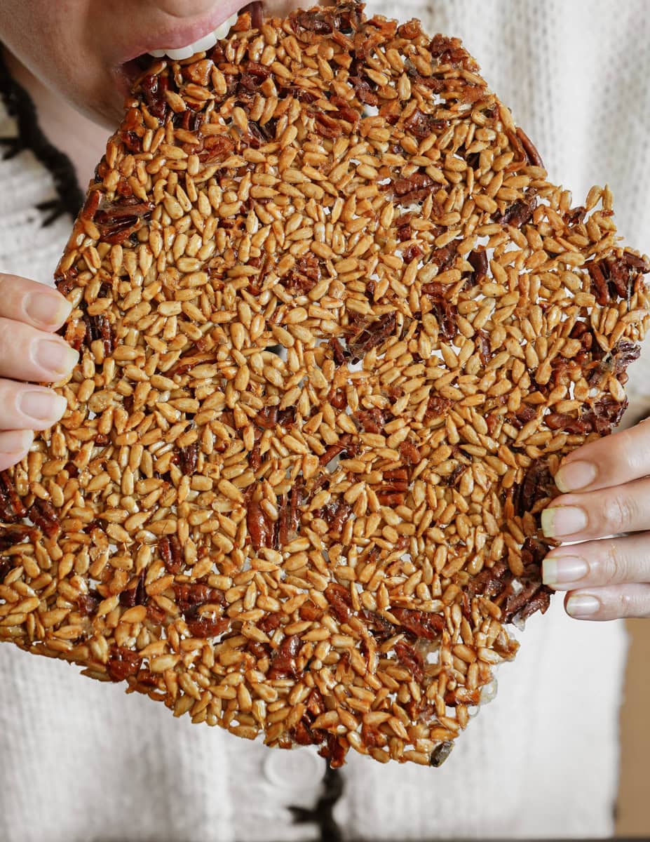 A person taking a bite of a large, square-shaped piece of caramelized nuts and seeds. The treat is glossy and densely packed with various grains and nuts, showcasing a textured and crunchy appearance.