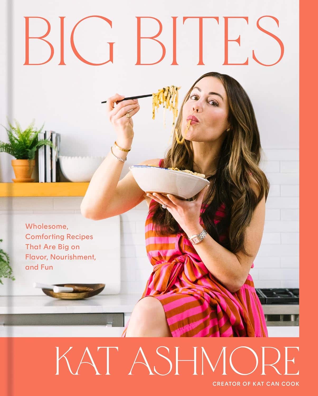 A woman with long hair eats a forkful of pasta while holding a bowl. She is seated in a kitchen environment with wooden shelves. The book cover reads Big Bites by Kat Ashmore, featuring recipes focused on flavor and nourishment.