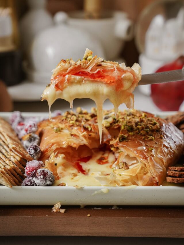 A knife holds a piece of baked brie wrapped in flaky pastry, topped with nuts and a red sauce. Cheese oozes from the center. Surrounding the dish are crackers and sugared cranberries, creating an inviting appetizer display.