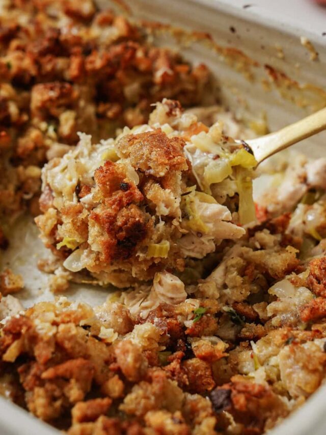 A close-up of a serving dish filled with a golden-brown stuffing, featuring chunks of bread, herbs, and vegetables. A spoon is scooping a portion from the dish, highlighting the dishs crispy and textured surface.