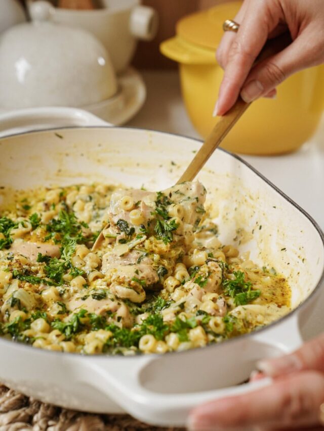 A person is serving pasta from a white baking dish using a wooden spoon. The dish contains creamy pasta with herbs and pieces of meat, garnished with fresh parsley. A yellow pot and kitchen items are in the background.