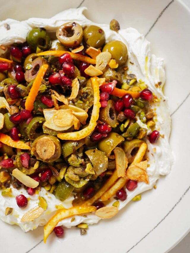 A vibrant dish featuring a bed of creamy spread topped with green olives, pomegranate seeds, crispy garlic slices, pistachios, and orange zest. Served on a white plate, this colorful mix showcases a variety of textures and flavors.