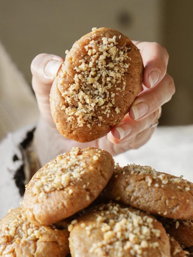 A hand holding a nut-topped cookie above a pile of similar cookies. The cookies are golden-brown and appear soft, with a sprinkling of chopped nuts on top. The background is softly blurred.