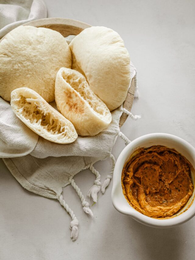 A wooden bowl with soft, fluffy pita bread on a cloth, next to a small white bowl filled with a creamy, spiced hummus dip on a light-colored surface.