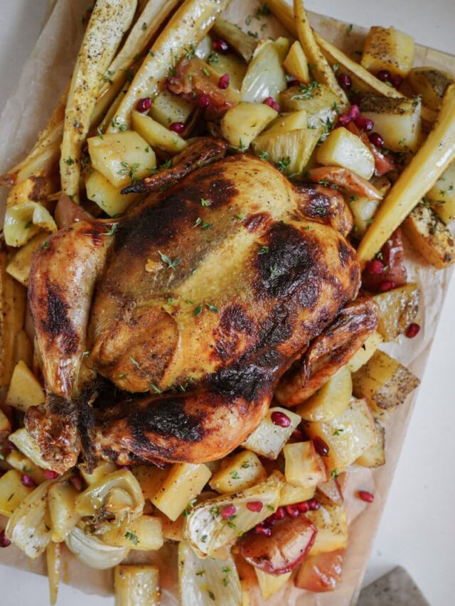 A roasted chicken surrounded by roasted potatoes, parsnips, and onions on a parchment-lined tray, garnished with herbs and pomegranate seeds.