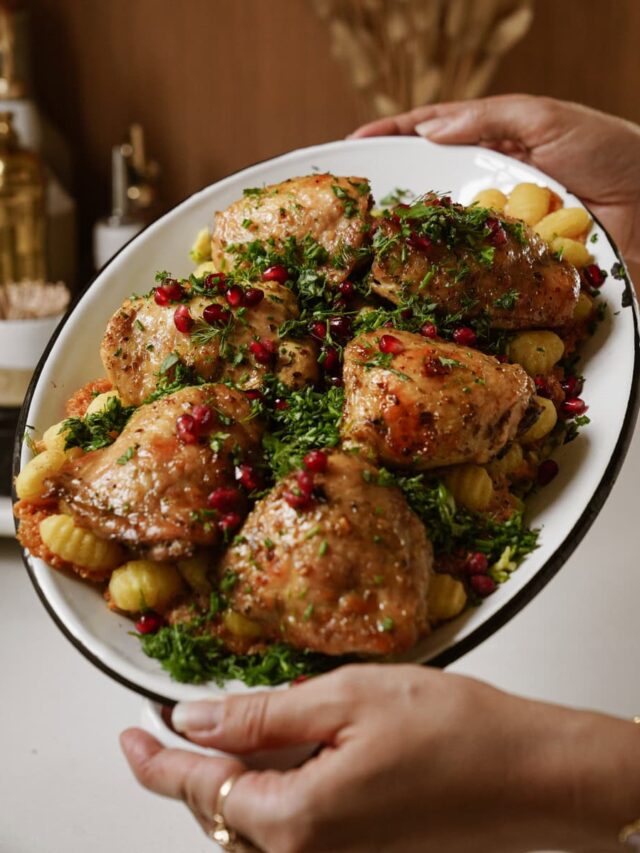 A platter of roasted chicken thighs garnished with chopped herbs and pomegranate seeds, served over roasted potatoes. The dish is being held up by two hands. The background is a blurred kitchen setting.