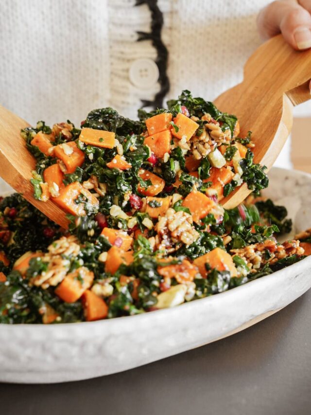 A person serves a colorful kale salad with sweet potatoes, pecans, and pomegranate seeds using wooden salad servers from a white bowl.