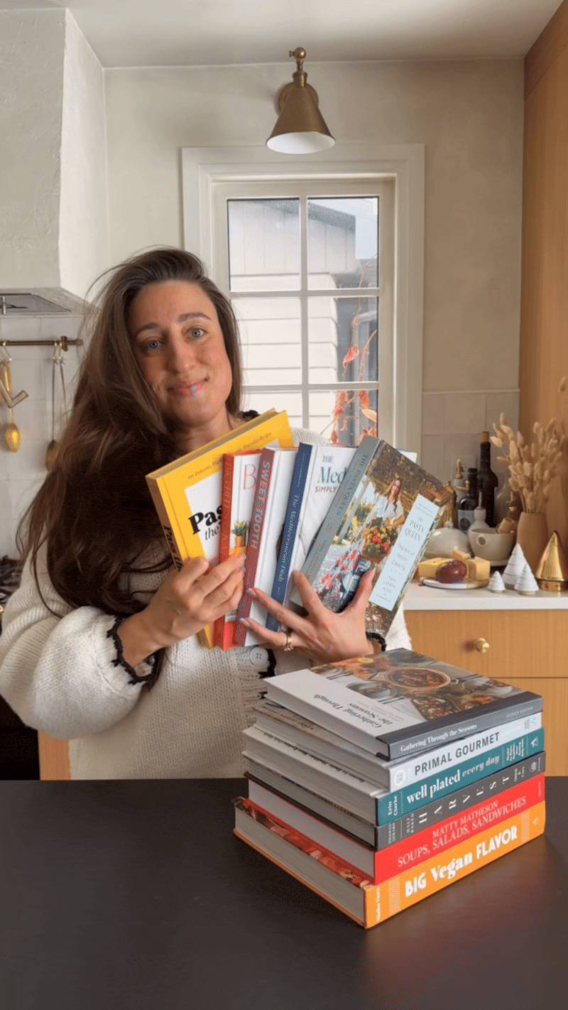 A woman in a kitchen holds several cookbooks while standing beside a counter with a stack of more cookbooks. The kitchen has a white tiled backsplash, wooden cabinets, and a window with a view of the outdoors.