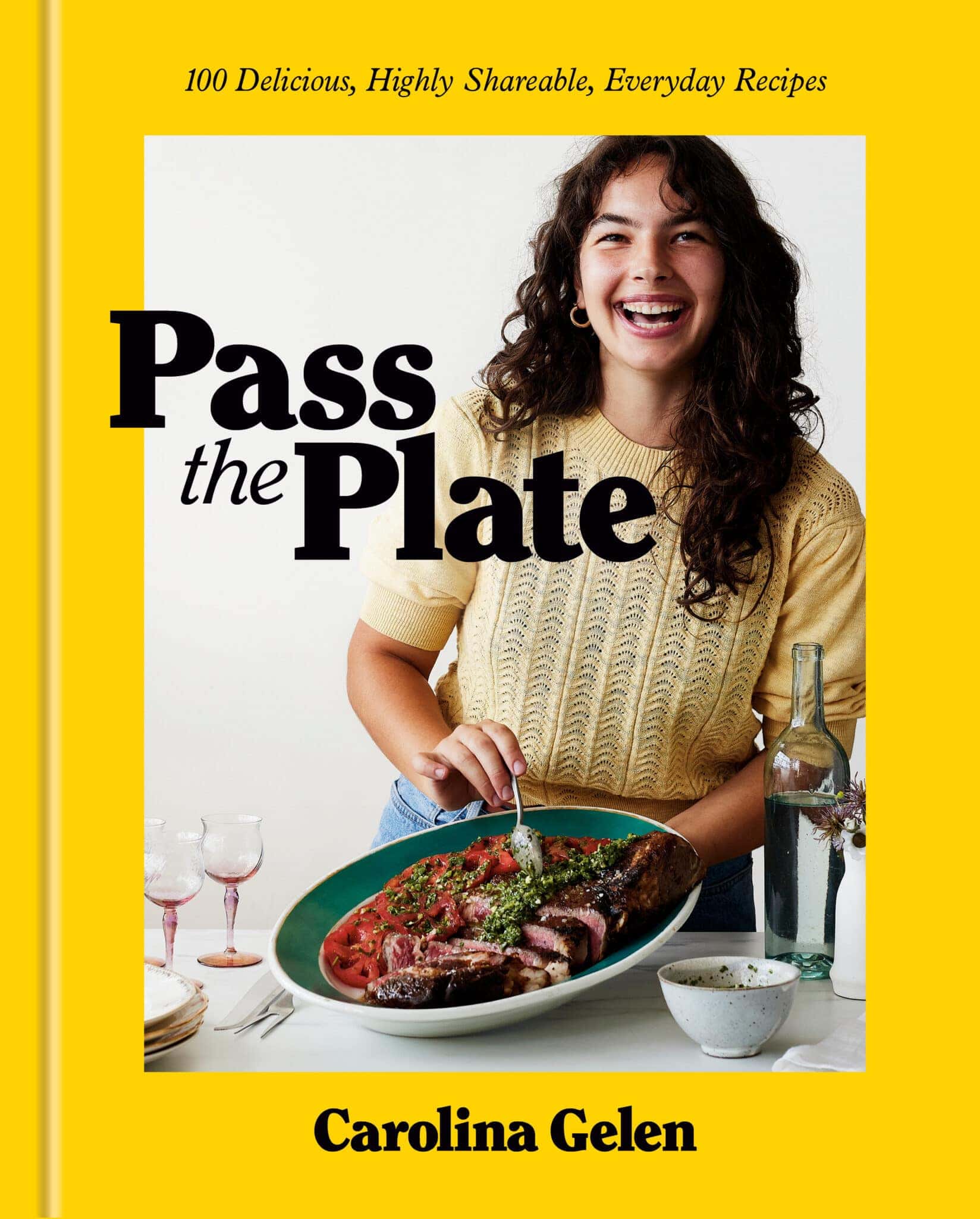 Cover of a cookbook titled Pass the Plate by Carolina Gelen. A person with long brown hair wearing a yellow sweater smiles and holds a platter of roasted meat. Wine glasses and a bottle are on the table. Bright yellow border.