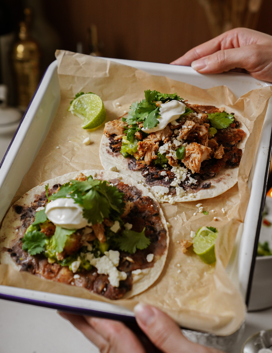 Two delicious tacos on a tray with parchment paper. Each taco is topped with meat, fresh cilantro, sour cream, black beans, and crumbled cheese. Lime wedges are on the side. Hands are holding the tray.