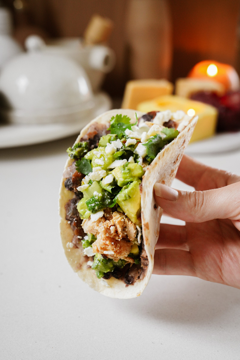 A hand holding a taco filled with black beans, avocado, cheese, cilantro, and crispy breaded pieces, set against a blurred background featuring a teapot and lit candles.